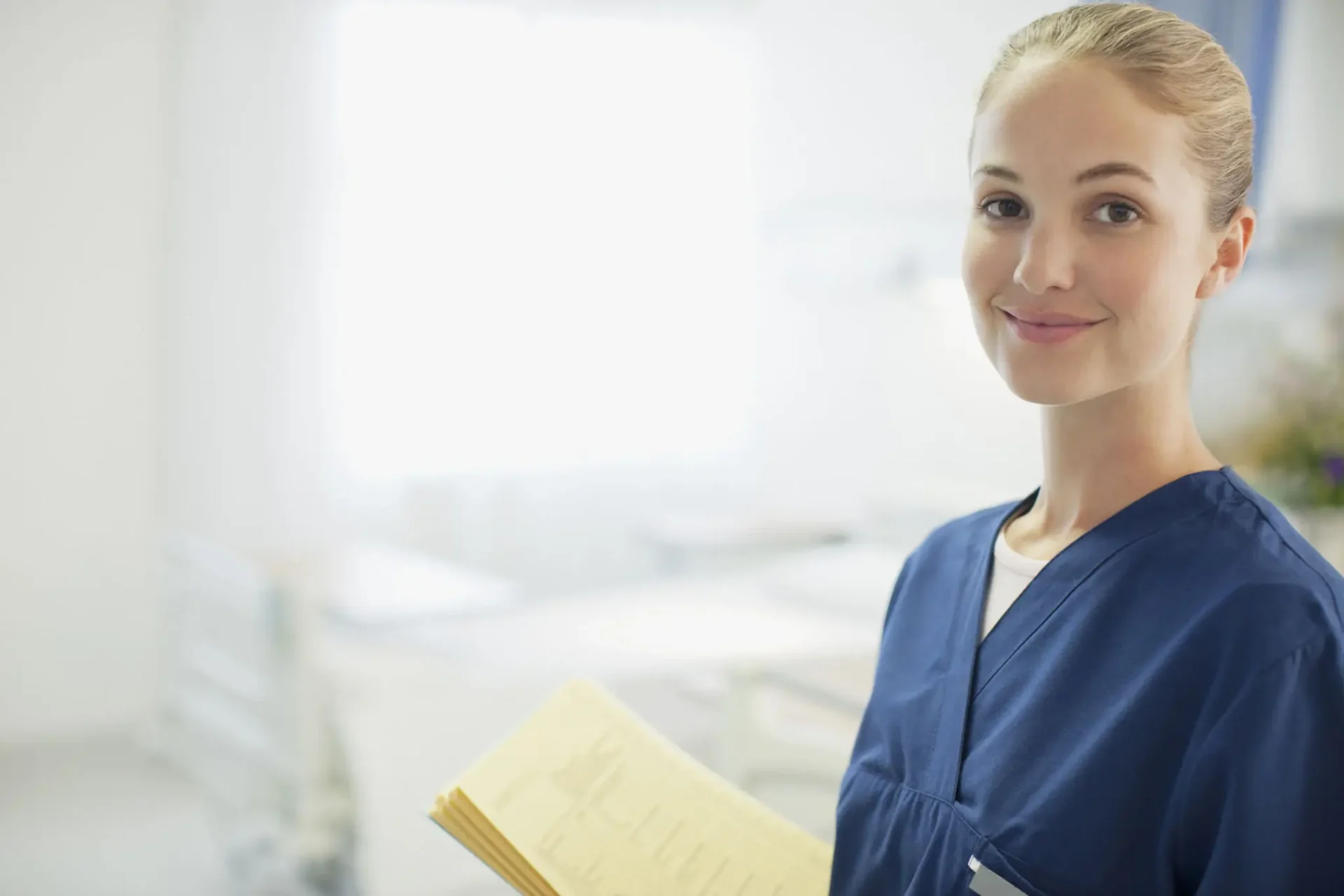 A nurse holding a clipboard, smiling gently at the camera in a bright medical setting.